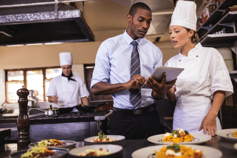 african american male manager holding tablet talking to chef inside large kitchen